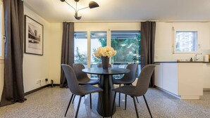 A dining area with a round table and four chairs, a vase with flowers, and a view of trees through large windows.