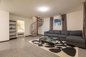 Ein modernes Wohnzimmer mit einem grauen Sofa, einem Glastisch, einer Wendeltreppe und einem Fenster mit Vorhängen.
