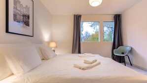 A hotel room with a large bed, a chair, a window with curtains, and a lamp.