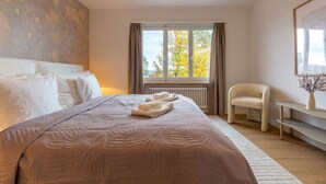 Quarto com cama grande, uma cadeira, mesa e janela com vista para árvores.