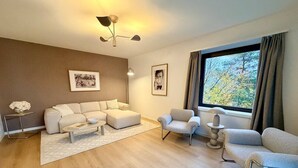Uma sala de estar moderna com um sofá, poltronas, uma mesa de centro e uma mesa lateral com abajur. Há quadros nas paredes, uma janela grande com cortinas e piso de madeira.