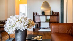 Un salon avec un canapé en cuir marron, une table basse noire sur laquelle repose un livre, et un vase rempli de fleurs blanches. On y trouve également une bibliothèque avec des livres et des objets décoratifs, ainsi qu’un miroir accroché au mur.