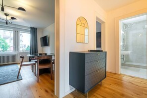 Una habitación moderna con piso de madera, un escritorio con silla, televisor, cómoda y zona de ducha.