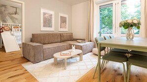 Een gezellige woonkamer met een bank, een kleine tafel en een eettafel met stoelen. Er hangen afbeeldingen in lijsten aan de muur en een raam met uitzicht op bomen.