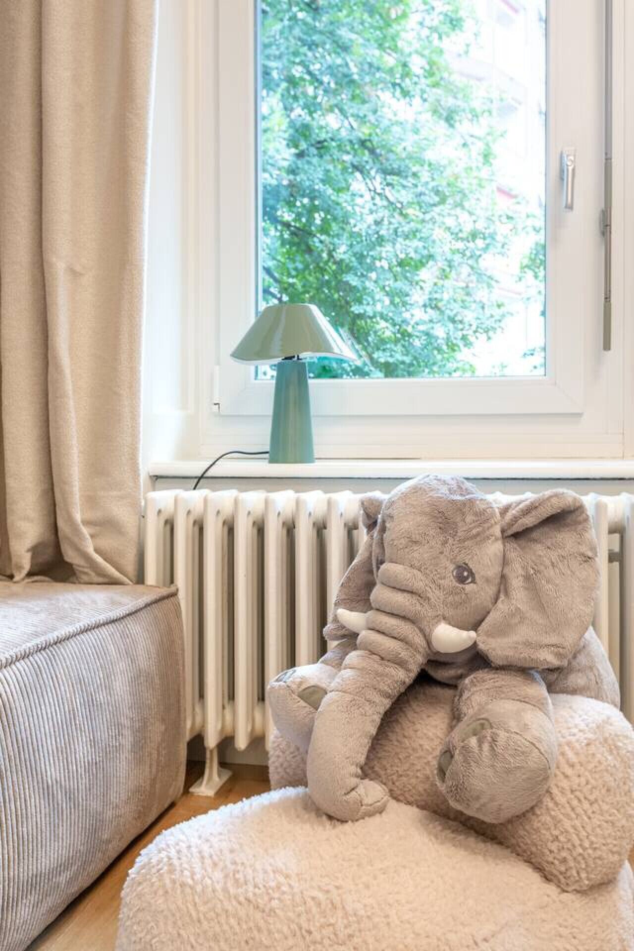 Tunnelmallinen huone, jossa on suuri ikkuna, patteri, lamppu ja pehmeä, elefantin muotoinen lelu.