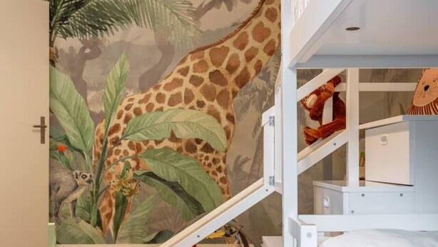 Een kinderspeelruimte met een muurschildering van een giraffe en begroeiing, witte meubels en een glijbaan.