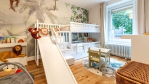 Een kinderkamer met stapelbedden, een glijbaan, een speelhoek met speelgoed en een groot raam met uitzicht op groen.