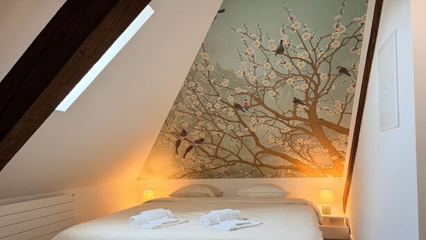 Une chambre avec un plafond en pente, un lit recouvert de draps blancs, deux lampes de chevet et une fresque murale représentant un arbre avec des oiseaux.