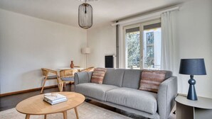 Un salon moderne avec un canapé gris, une table basse ronde en bois, un coin repas avec des chaises en osier et une grande fenêtre avec des rideaux.