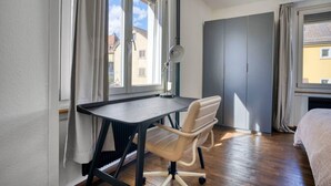 Une chambre à coucher avec un lit, un bureau et une chaise. Il y a des fenêtres avec des rideaux, un radiateur et une armoire.