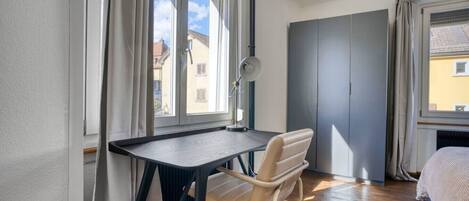 Un dormitorio con una cama, un escritorio y una silla. Hay ventanas con cortinas, un radiador y un armario.