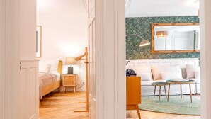 En hall leder till ett sovrum med en säng, ett sängbord och en lampa. Ett vardagsrum med en soffa, ett soffbord och stolar syns genom en öppen dörr.