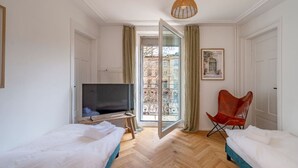 Ett hotellrum med två sängar, en tv, en röd fåtölj och ett stort fönster med utsikt över byggnader.