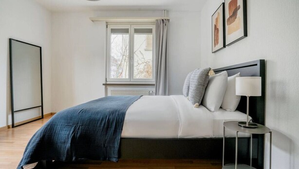 Ein modernes Schlafzimmer mit Bett, Nachttisch, Lampe, Fenster und einem gerahmten Bild an der Wand.
