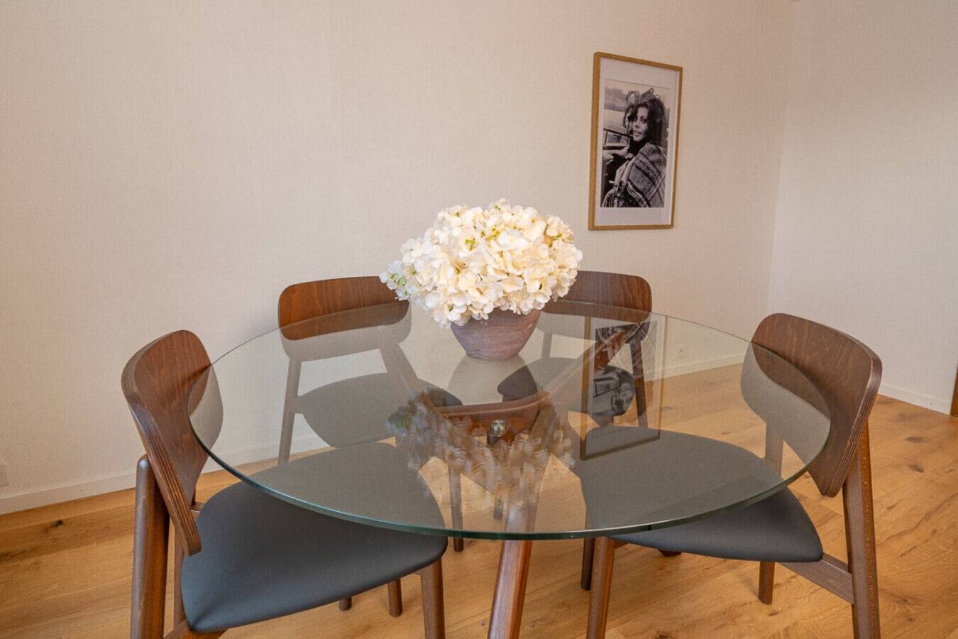 Une table ronde en verre, accompagnée de quatre chaises en bois et d’un vase rempli de fleurs blanches.