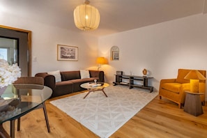 Una sala moderna con un sofá marrón, una mesa de centro de vidrio, una mesita auxiliar redonda, una lámpara amarilla y piso de madera.
