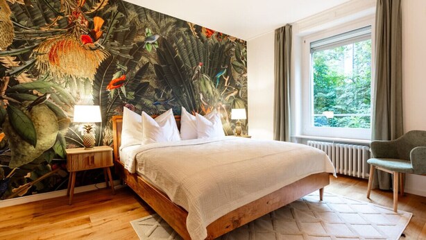 A bedroom with a large mural of tropical birds and foliage, a wooden bed with white linens, a nightstand with a lamp, and a view of greenery through a window. - Stylish 3-BR design flat with terrace · Kreis 6 (Zürich)