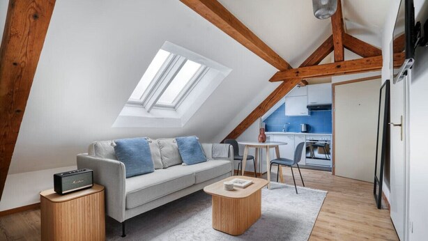 Un appartement moderne de style « loft » avec un plafond en pente, un canapé gris agrémenté de coussins bleus, une table basse en bois, une petite table d’appoint avec une radio, et une vue sur la cuisine à travers une porte ouverte.