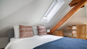Une chambre avec un plafond en pente, un puits de lumière, un lit avec une literie blanche et bleue, et des meubles en bois.