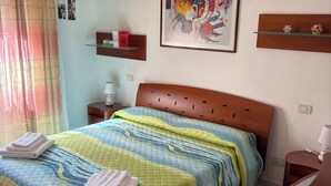 A neatly made bed with a striped bedspread and folded towels, a wooden headboard, a bedside table with a lamp, a framed abstract painting on the wall, and a small shelf with decorative items. - Poetto Casa Bach Ck, Cozy apartment a few steps from the sea (Quartu Sant'Elena)