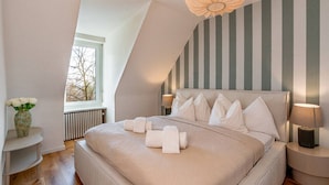 Une chambre moderne avec un plafond en pente, un grand lit agrémenté de plusieurs oreillers, des lampes de chevet et un vase de fleurs posé sur une table d’appoint.