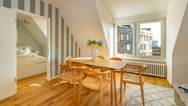 Une pièce chaleureuse avec une table à manger en bois, des chaises et un vase rempli de fleurs. On y trouve un lit avec une literie blanche et, par la fenêtre, une vue sur les bâtiments environnants.