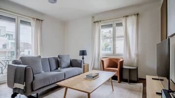 Un salon moderne avec un canapé gris, une table basse en bois, un petit fauteuil orange et un téléviseur posé sur un meuble.