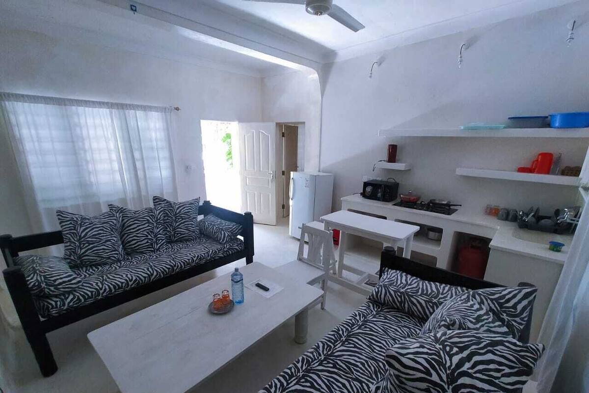 Un espace de vie compact comprenant un canapé à motifs zébrés, une petite table à manger, un coin cuisine et une porte donnant sur un espace extérieur.