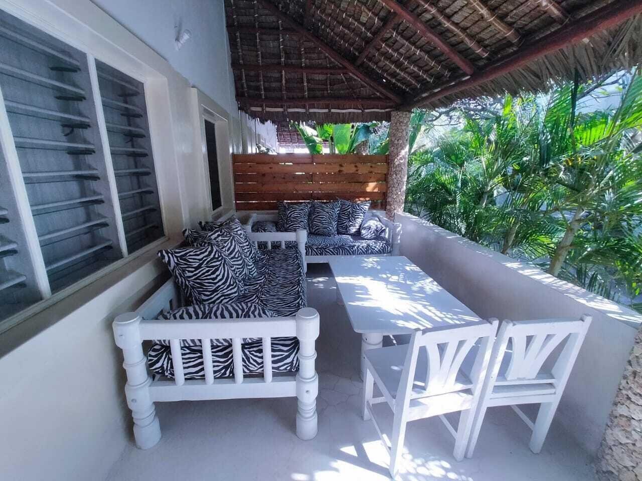 A covered outdoor seating area with a wooden bench and a table, surrounded by lush greenery.