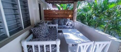 A covered outdoor seating area with a wooden bench and a table, surrounded by lush greenery.