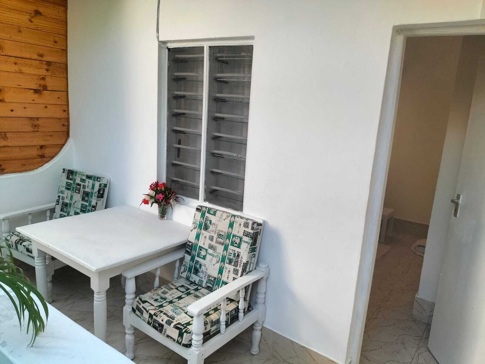 A white dining area with a table and two chairs, a window with blinds, and a door leading to another room.