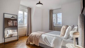 A bedroom with a bed, a bedside table, a lamp, a mirror, and a window with curtains. - Bright 3-BR in Wipkingen — rooftop views, free parking & garden (Zürich)