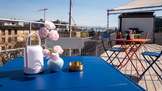 Une terrasse sur le toit avec une table bleue, un vase blanc orné de fleurs roses et un bol en métal.
