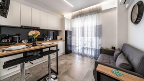 Una cocina moderna con armarios blancos, un comedor con mesa de madera y sillas negras, y un sofá gris en una sala de estar con vistas a través de cortinas translúcidas.