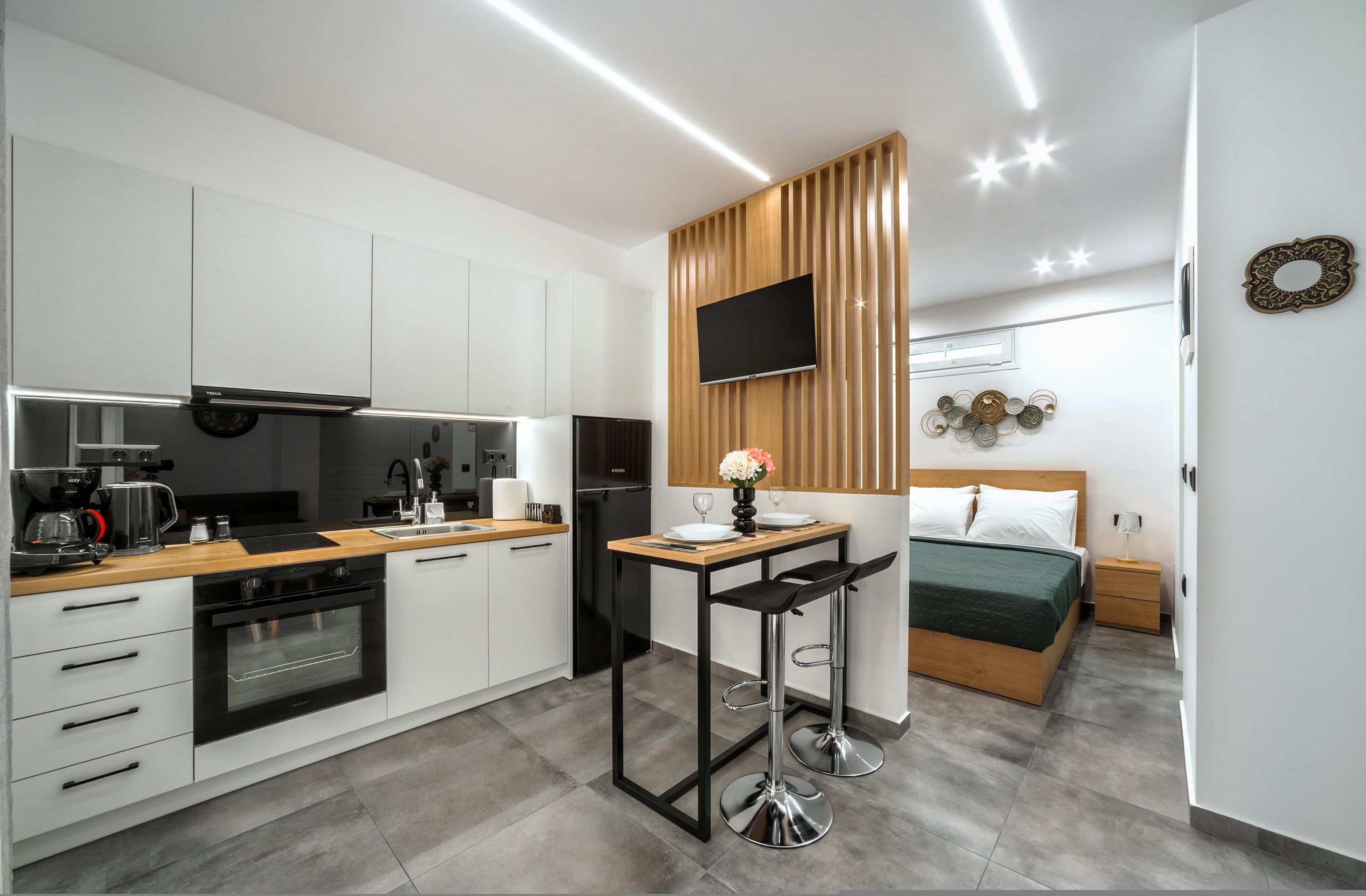 Een moderne keuken met witte kasten, een houten aanrechtblad en een inbouwoven. Er is een eettafel met twee stoelen en een televisie aan de muur bevestigd.