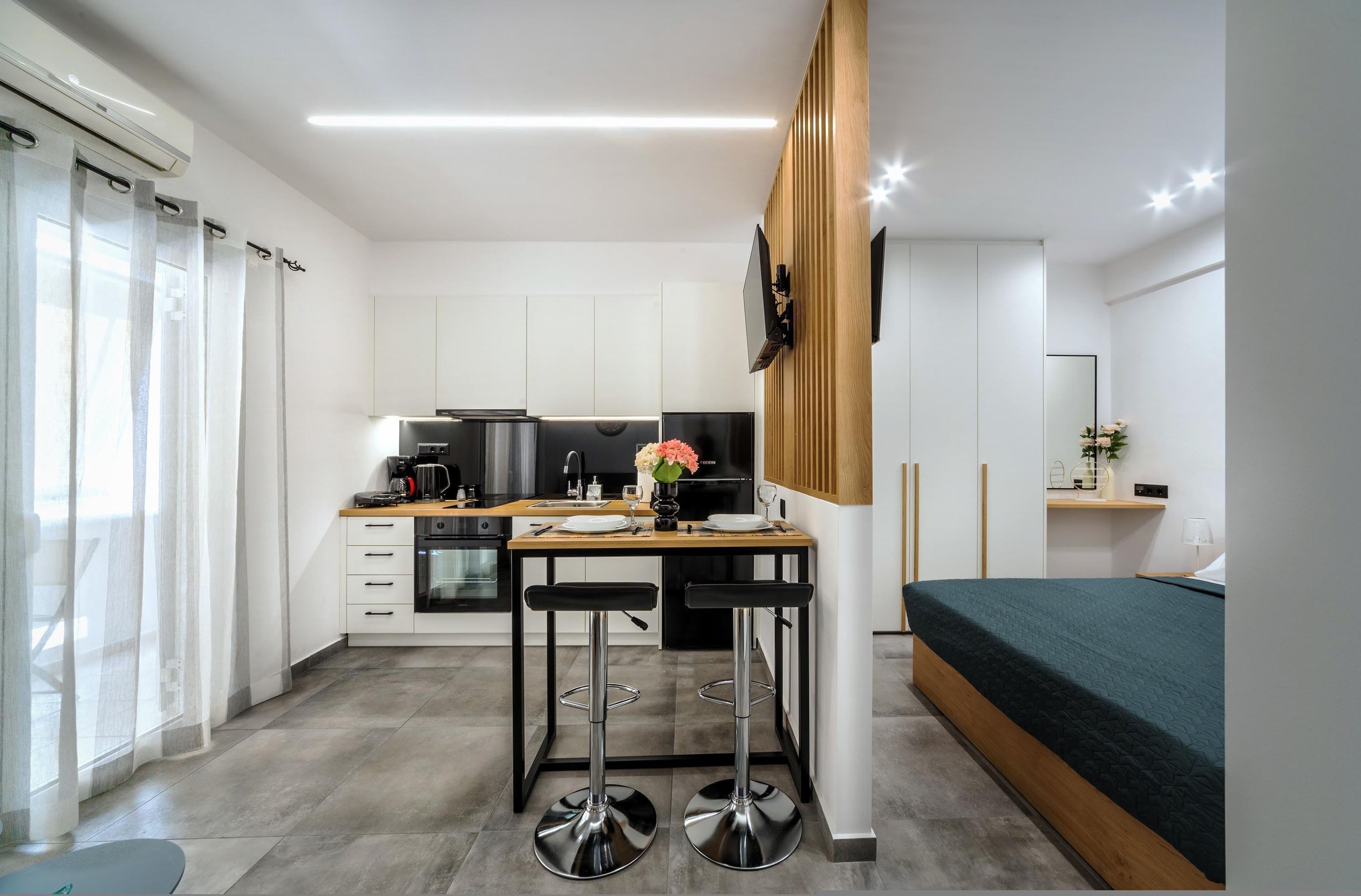 A modern kitchen with a dining area, a TV mounted on the wall, and a bed with a green bedspread.