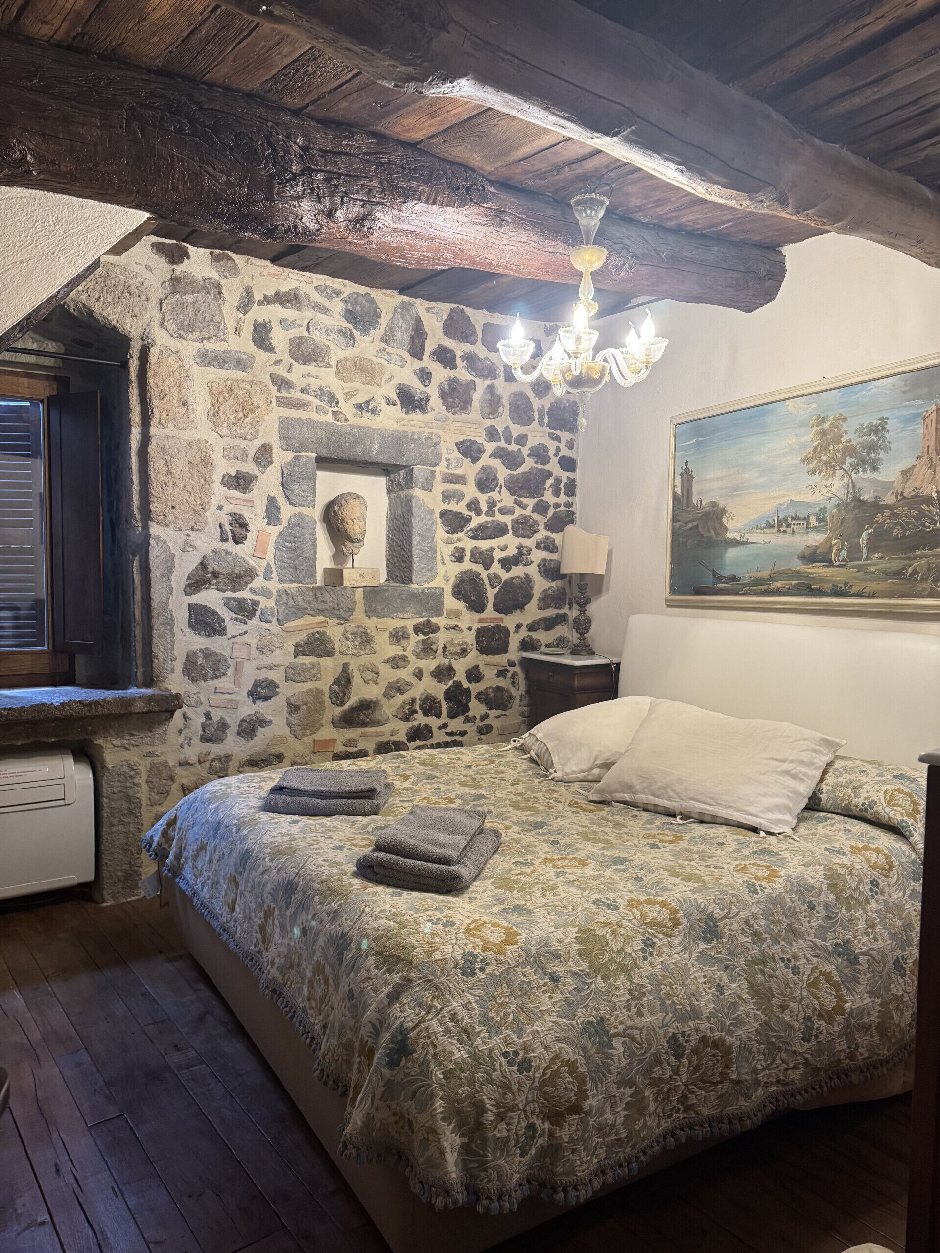 A bedroom with a stone wall, a bed with a floral bedspread, a nightstand with a lamp, and a painting on the wall.