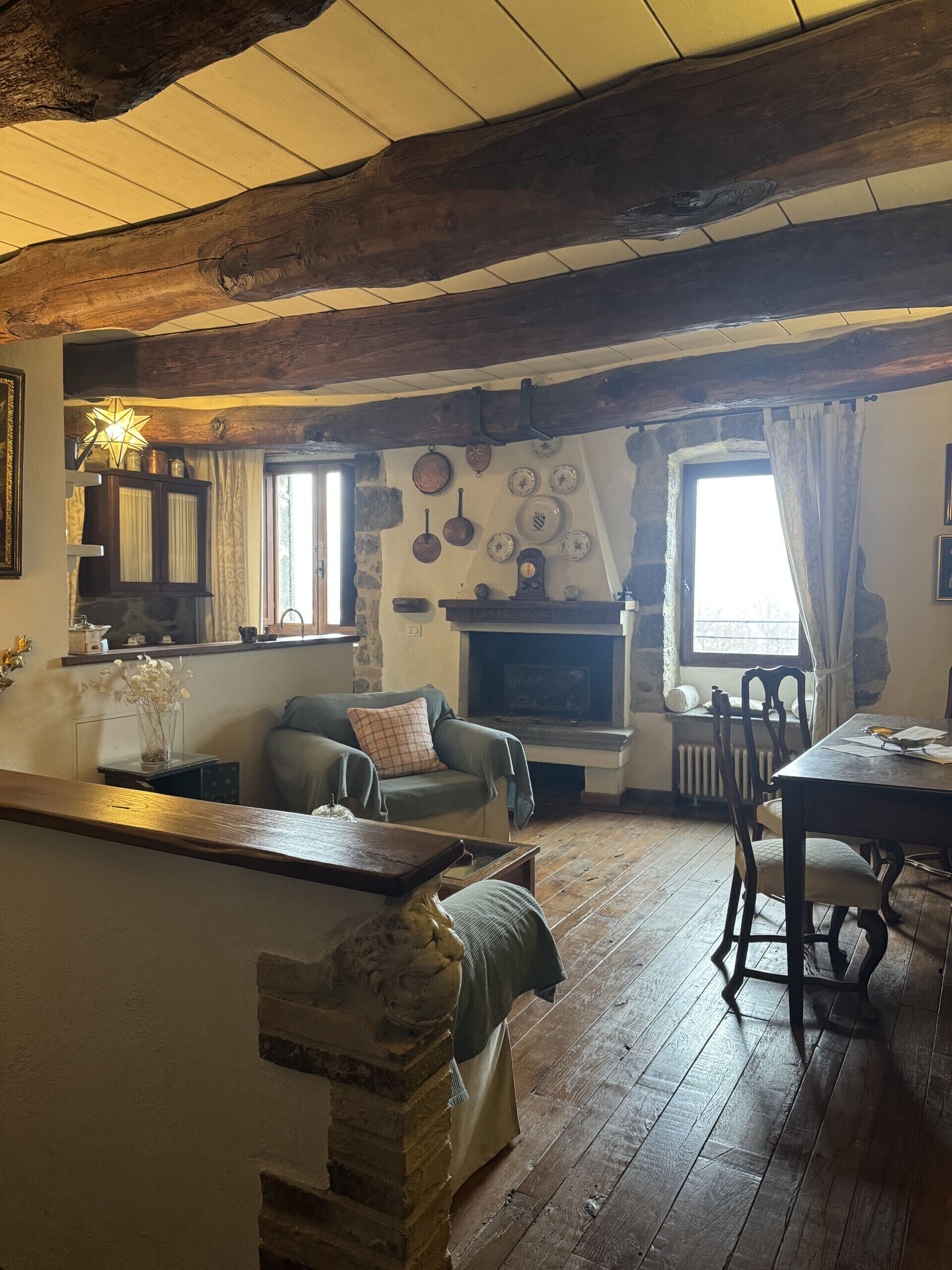 Een rustieke woonkamer met zichtbare houten balken, een stenen openhaard en een eethoek met een houten tafel en stoelen.
