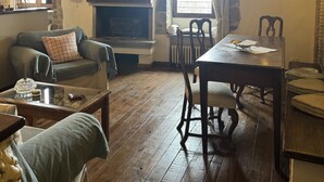 A cozy living room with a fireplace, wooden beams, a stone wall, and a dining area with a table and chairs.