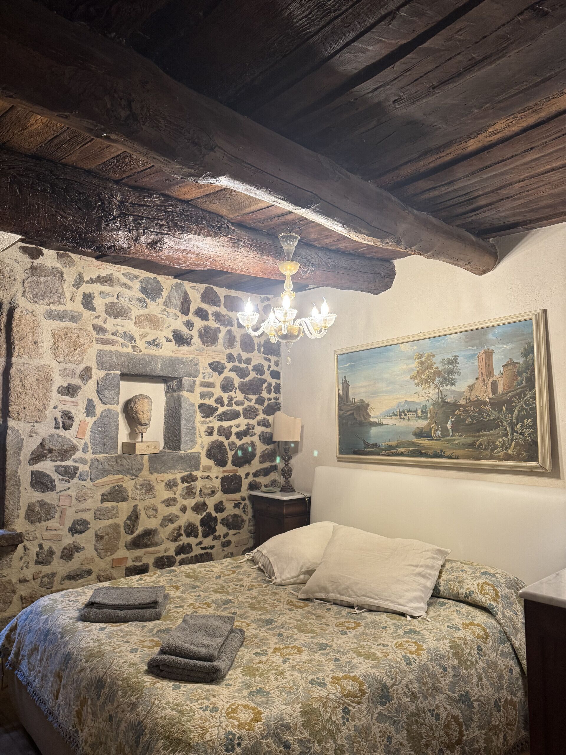 Une chambre avec un mur en pierre, un lit recouvert d’une literie à motifs floraux, un lustre et un tableau représentant un paysage.