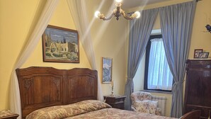 Een slaapkamer met een houten bed, nachtkastje, dressoir, raam met gordijnen en een kroonluchter.