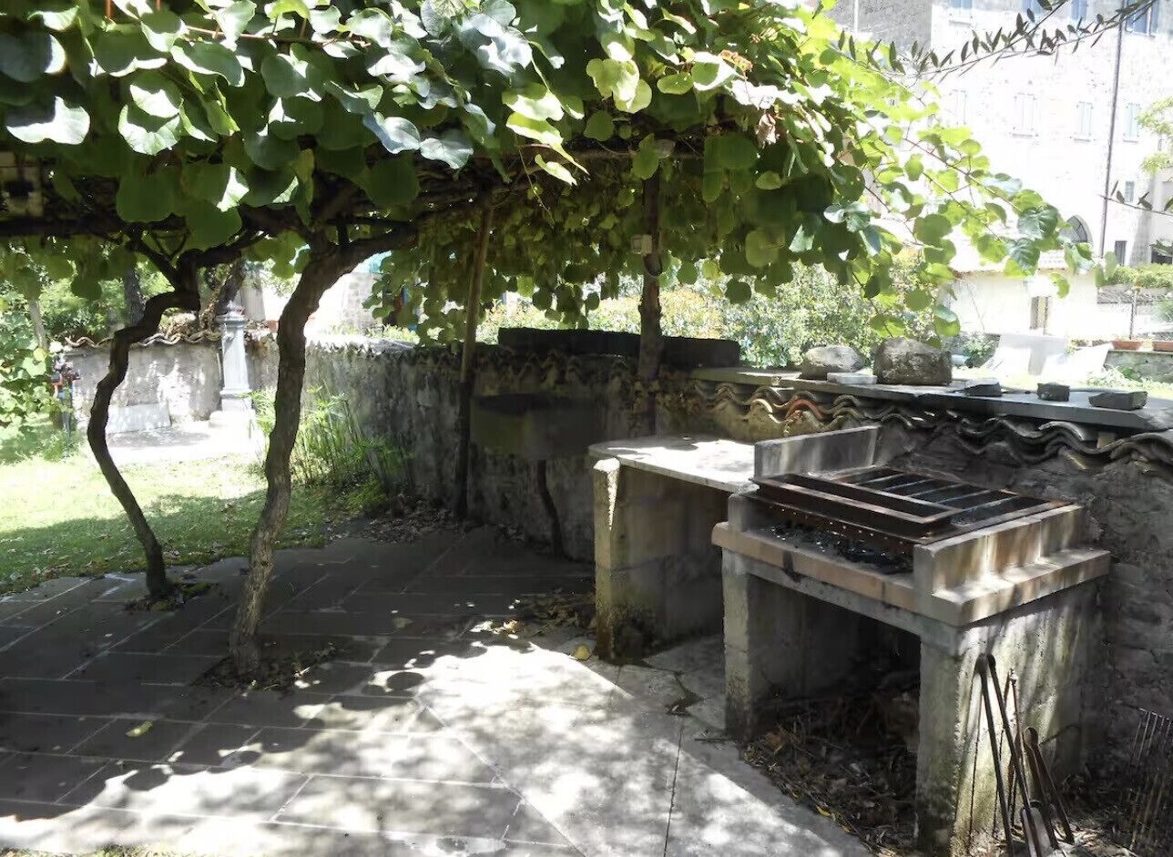 An outdoor area with a stone bench, a grill, and a tree providing shade.