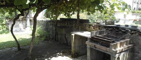 An outdoor area with a stone bench, a grill, and a tree providing shade.