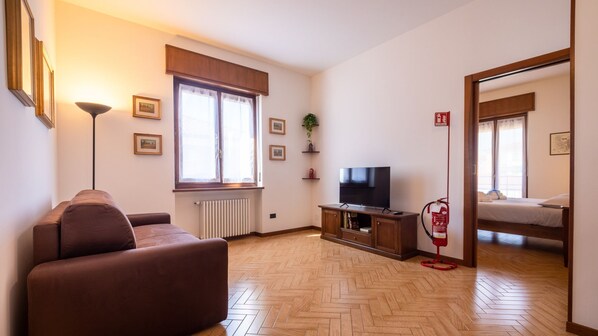 A furnished living room with a sofa, a television, a radiator, a window with curtains, and a fire extinguisher. - Appartamento  Marullina (Verona)