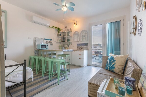 A compact living space with a kitchenette, a dining area with green stools, a sofa with a tray of candles, and a view through a glass door.