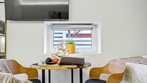 Moderni huone, jossa on pyöreä pöytä, kaksi puutuolia, seinälle asennettu televisio ja ikkuna, josta on näkymä ulkona olevaan rakennukseen.