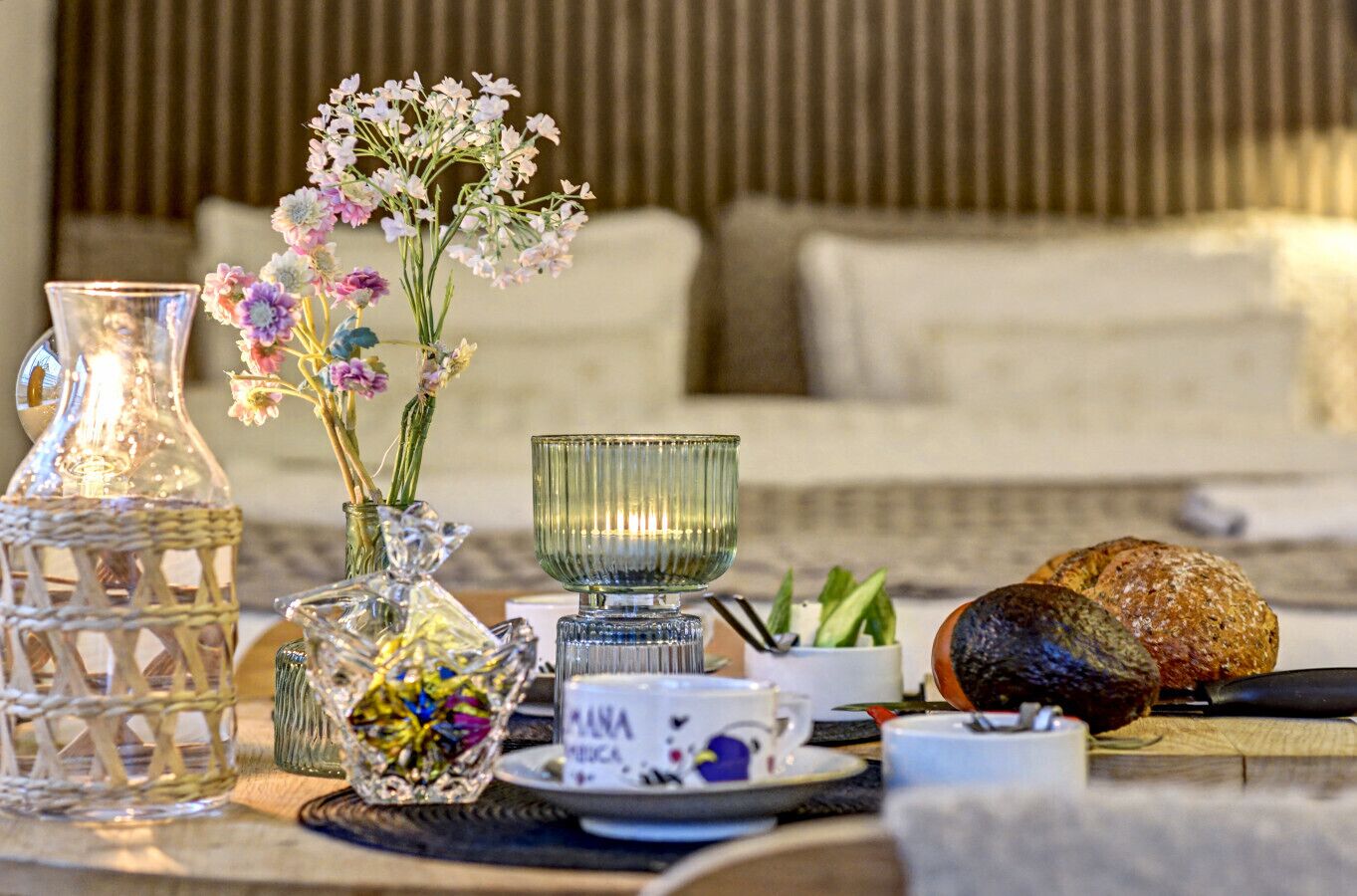 A table set with a clear glass pitcher, a glass bowl, a vase with flowers, a plate with a cup, and a loaf of bread.