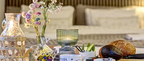 A table set with a clear glass pitcher, a glass bowl, a vase with flowers, a plate with a cup, and a loaf of bread.