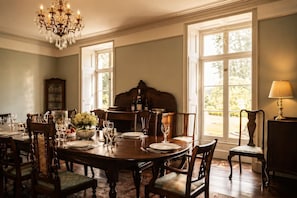 A dining room with a chandelier, large windows, a piano, and a lamp.