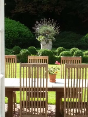 Une table en bois, quatre chaises, une plante en pot et une vue sur un jardin bien entretenu, agrémenté de haies et d’un grand jardinière.